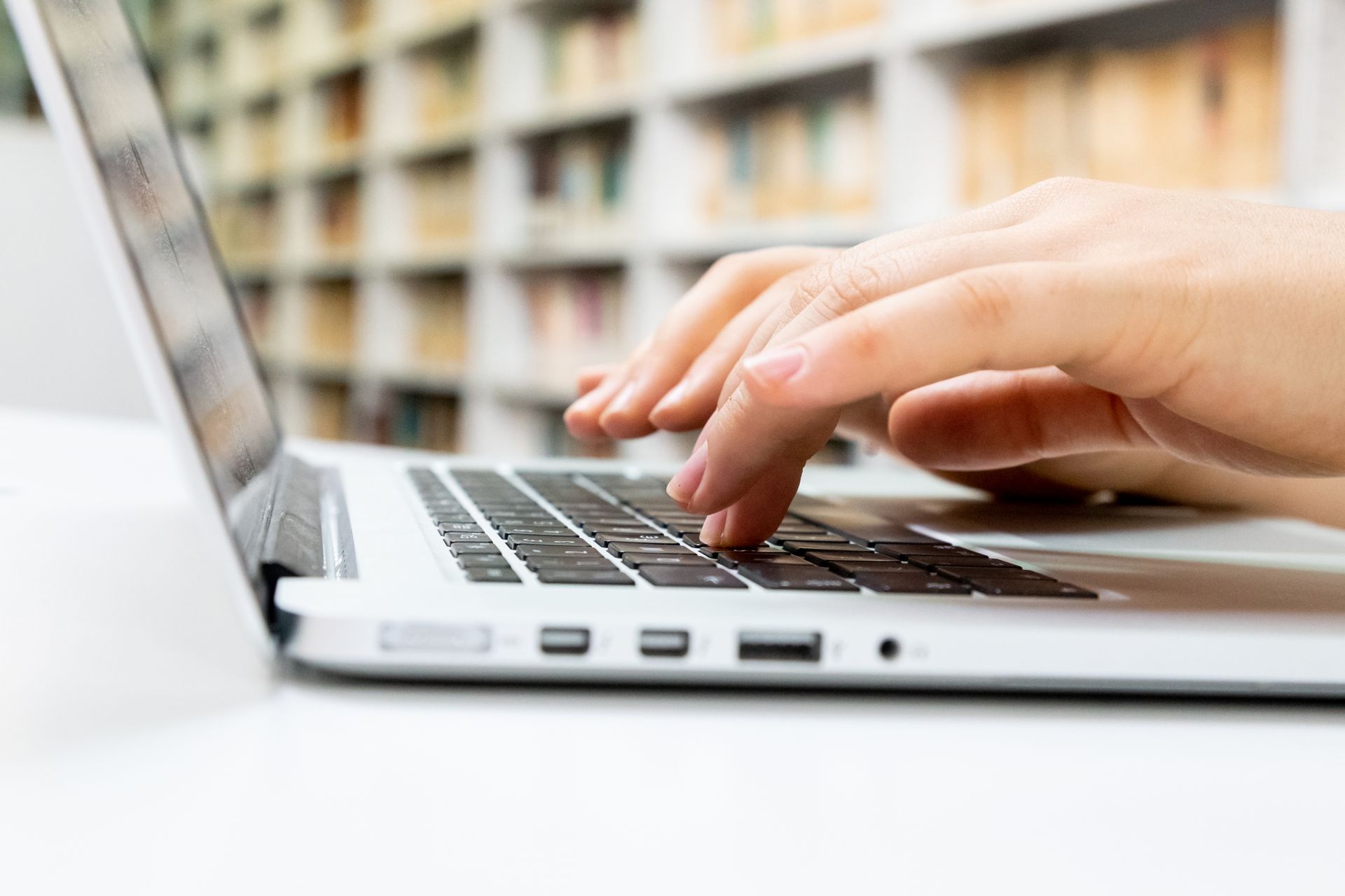 Hands typing on a laptop keyboard with bookshelves in the background.