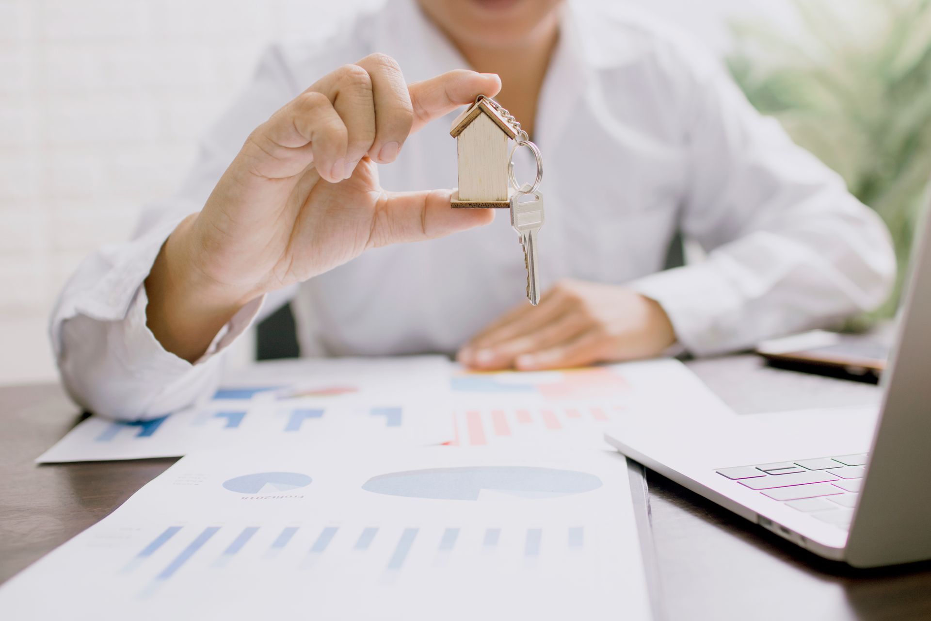 A person holds a key with a wooden house keychain above papers beside a laptop.