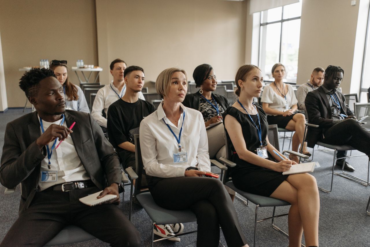 People wearing badges sit in rows of chairs, with several holding notebooks and pens.