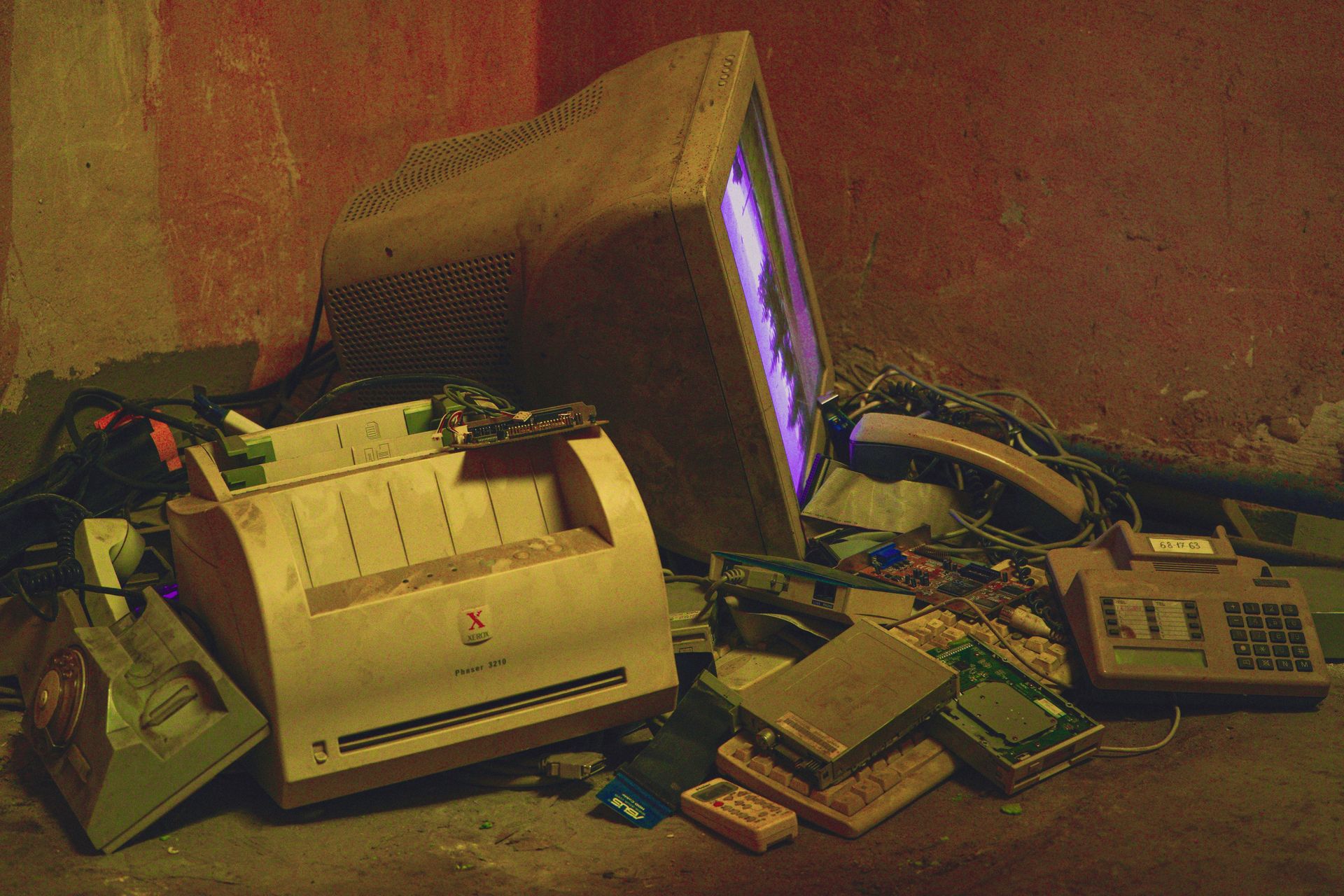 An old television, printer, telephone and dismantled electronics piled against a wall.