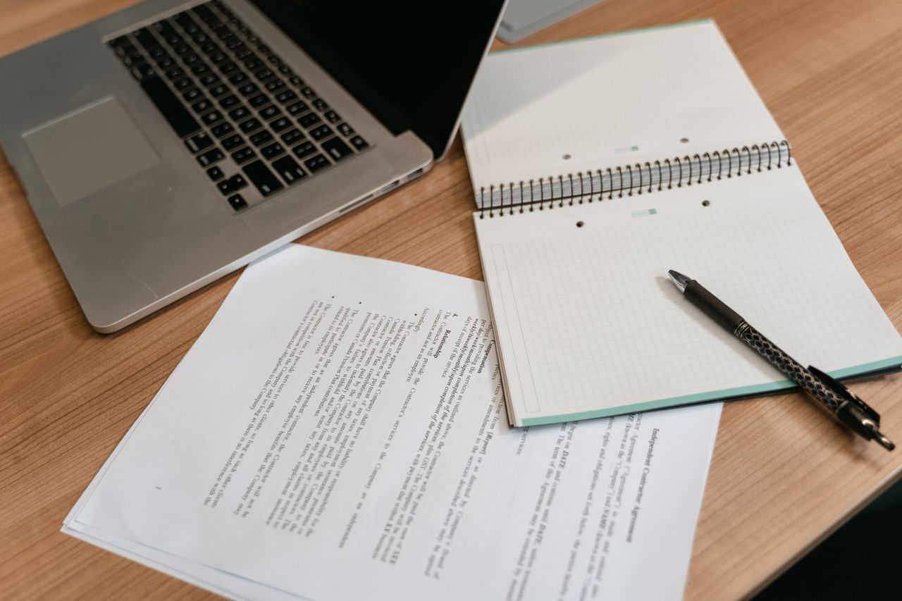 A laptop, spiral notebook, pen and printed papers on a wooden desk.