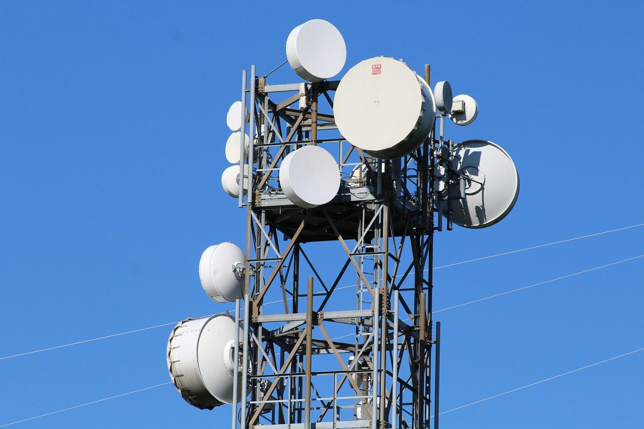 A metal communications tower supports several round white antenna dishes beneath a blue sky.