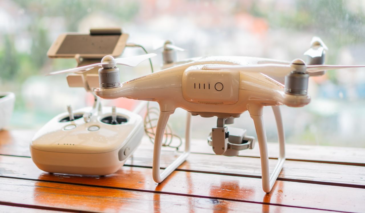 A white quadcopter with a camera underneath sits on a wooden table beside its remote controller.