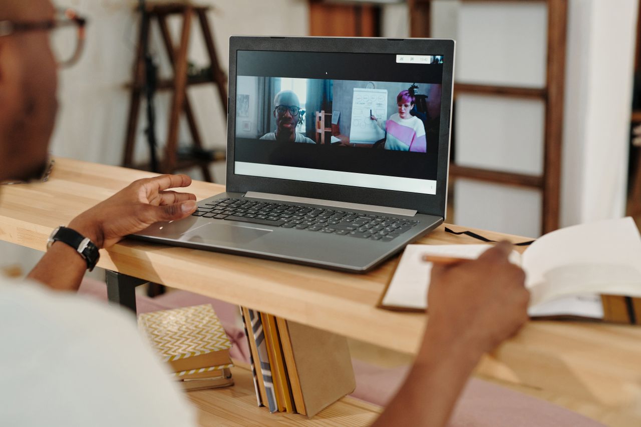 A person holds a pen beside a notebook while a laptop displays two people on a video call.