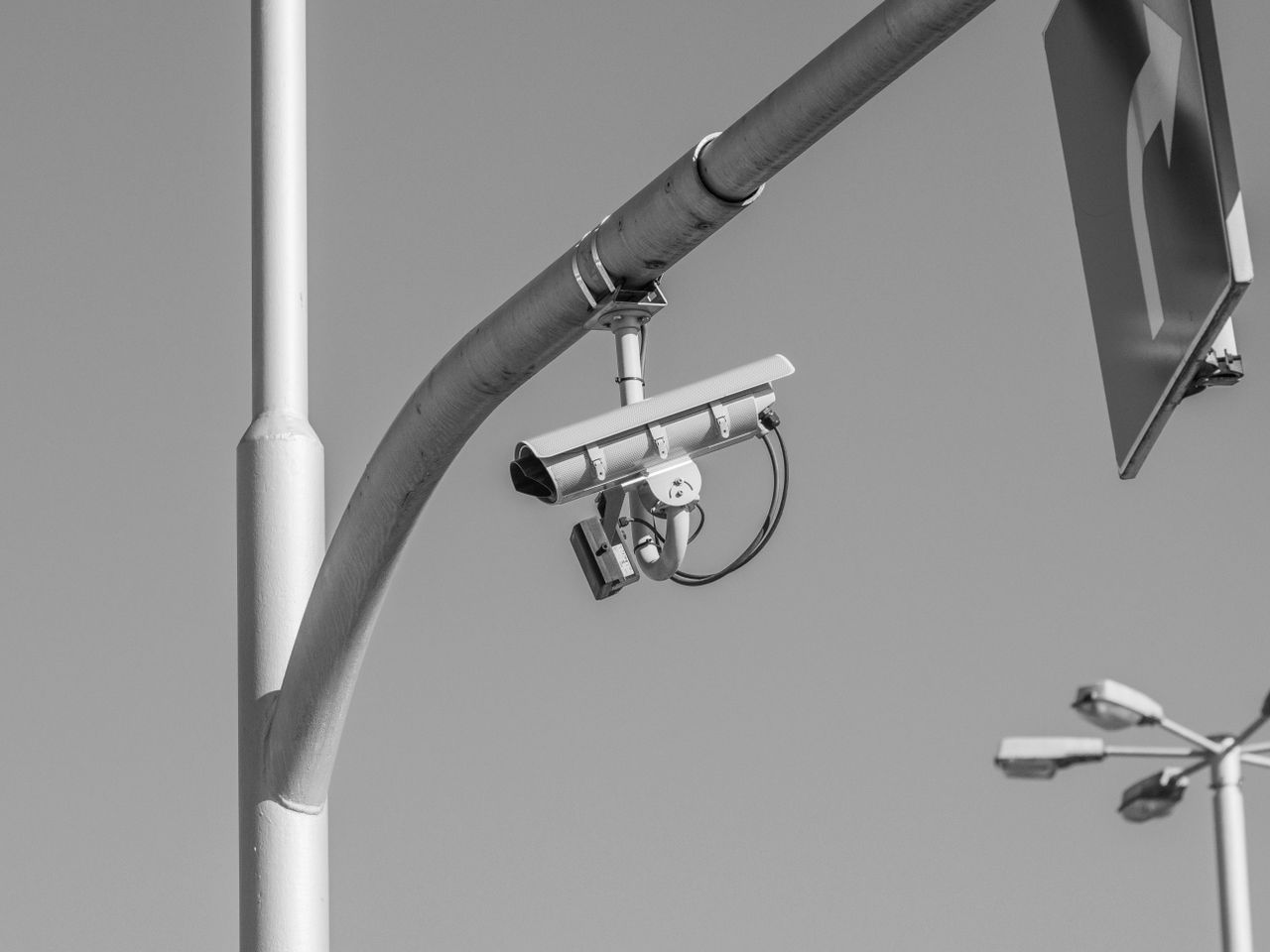 A surveillance camera hangs from a curved streetlight pole in a black and white photograph.