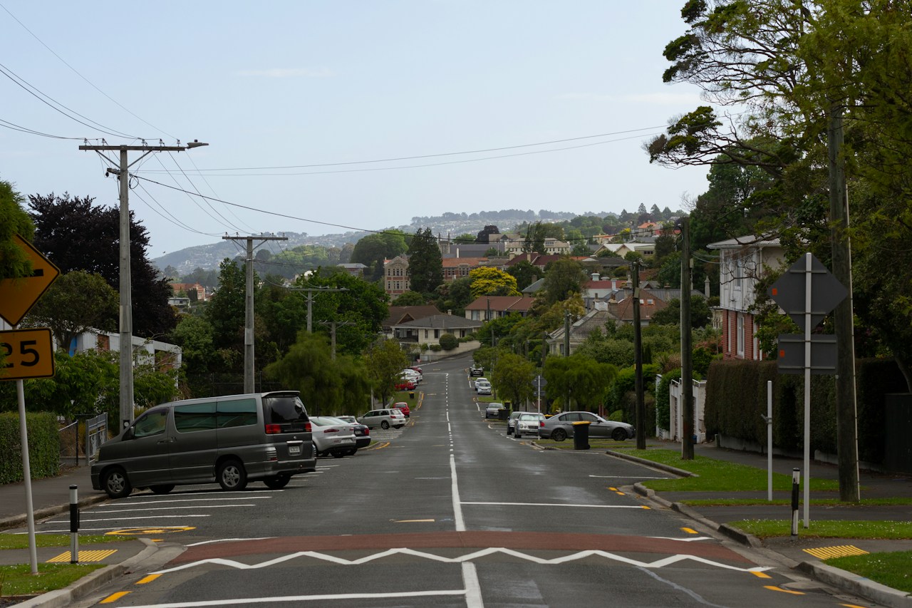 Parked cars line a sloping residential street with homes, trees, utility poles and painted crossing markings.