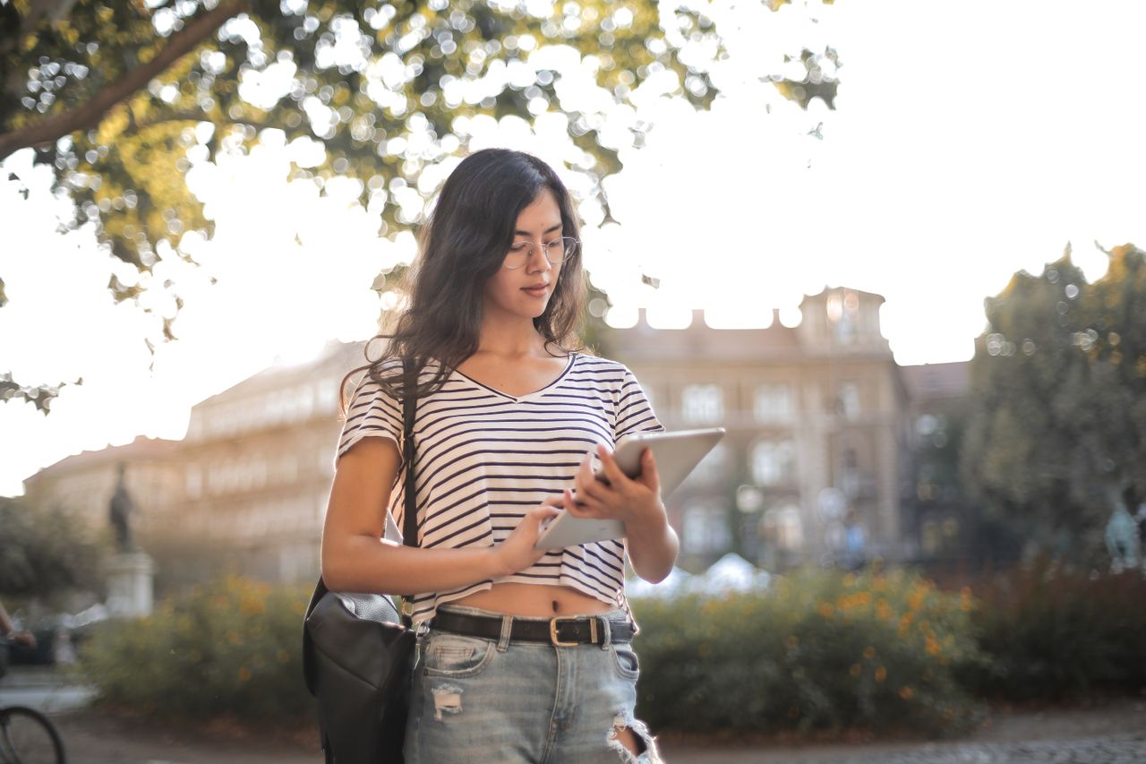 A woman wearing glasses and a striped shirt uses a tablet outdoors beneath leafy trees.