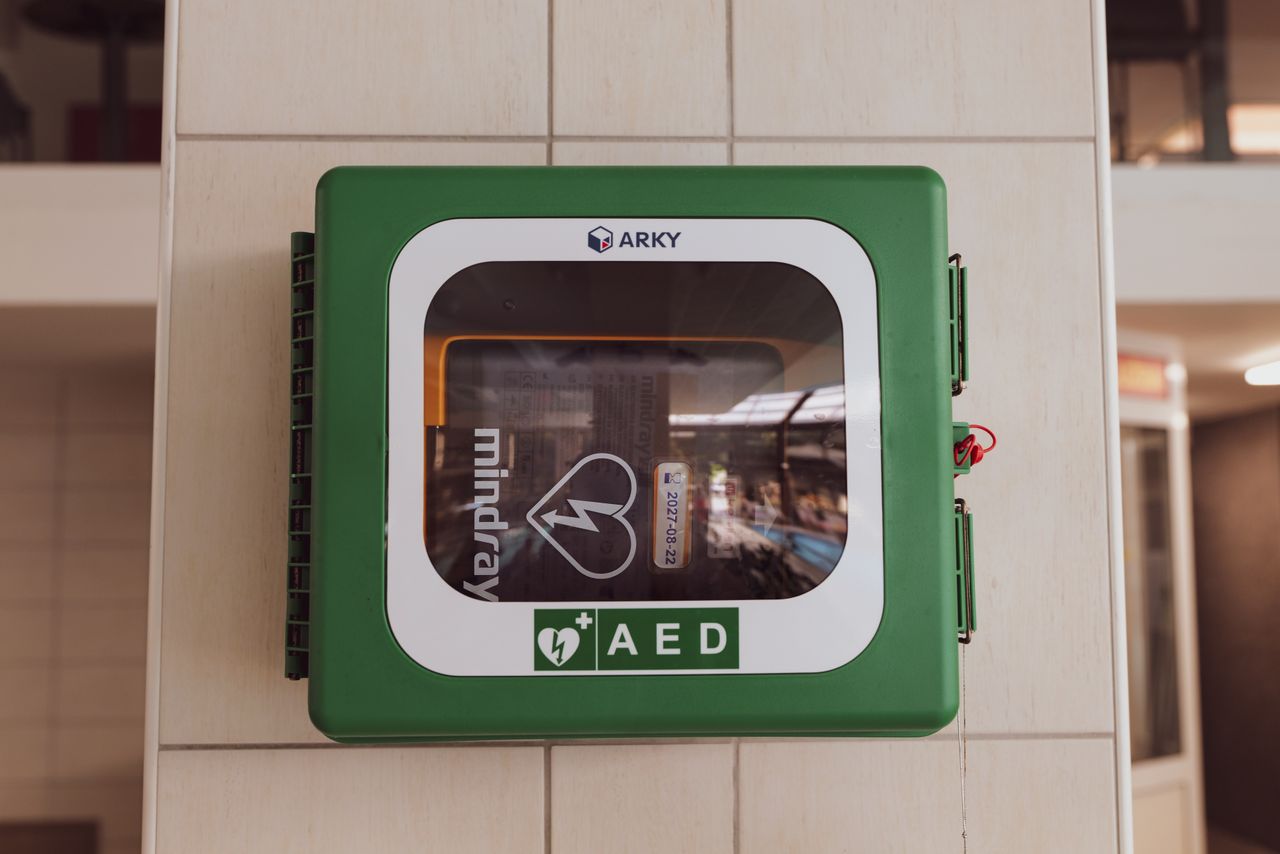 An AED inside a green cabinet with a clear front panel, mounted on a tiled indoor wall.