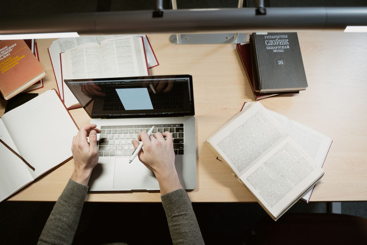 An overhead view of a person using a laptop and holding a pen beside open books on a desk.