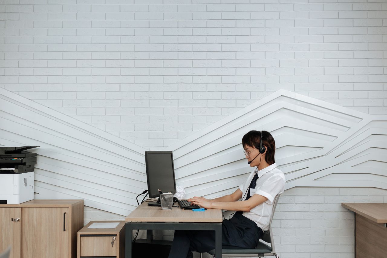 A woman wearing a headset works at a desktop computer beside a printer in an office.