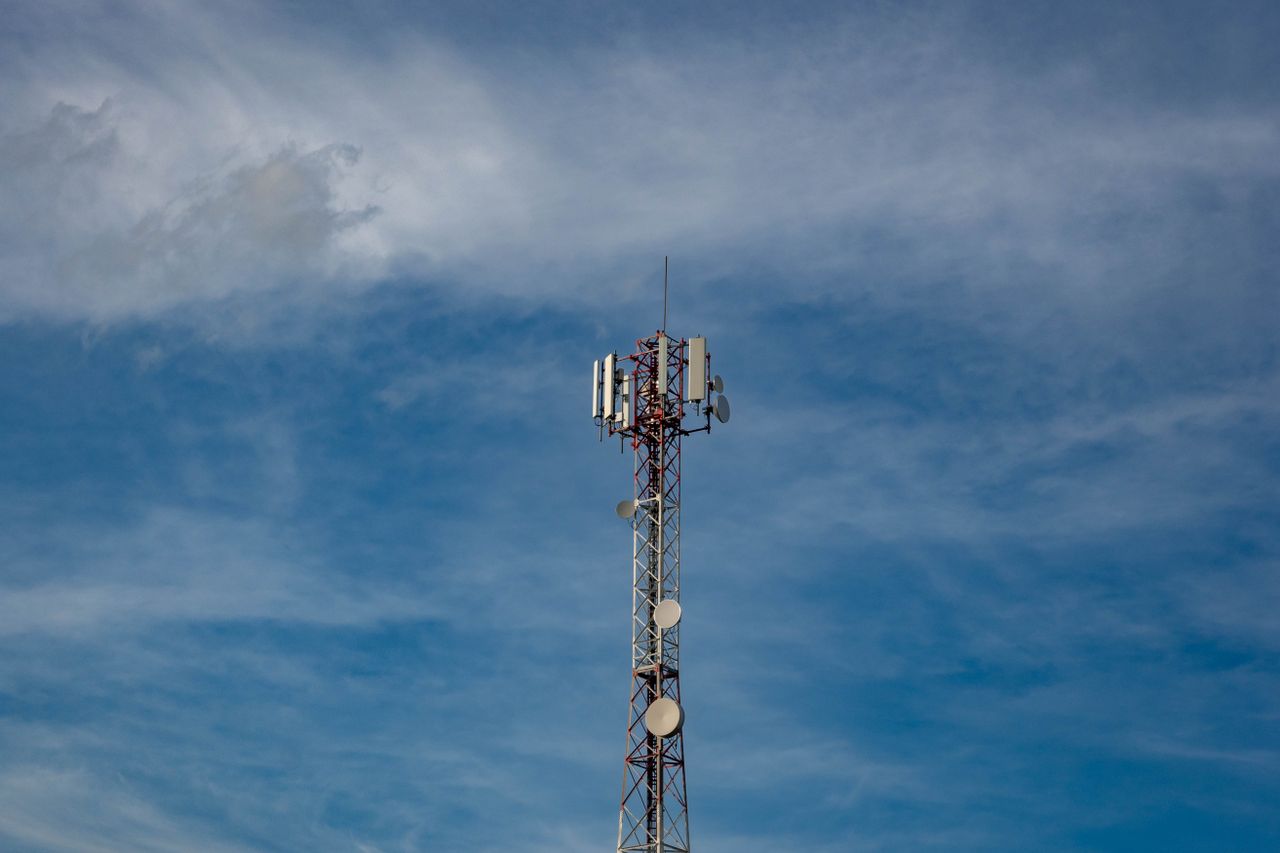 A metal communications tower with panel antennas and circular dishes rises beneath a cloudy blue sky.