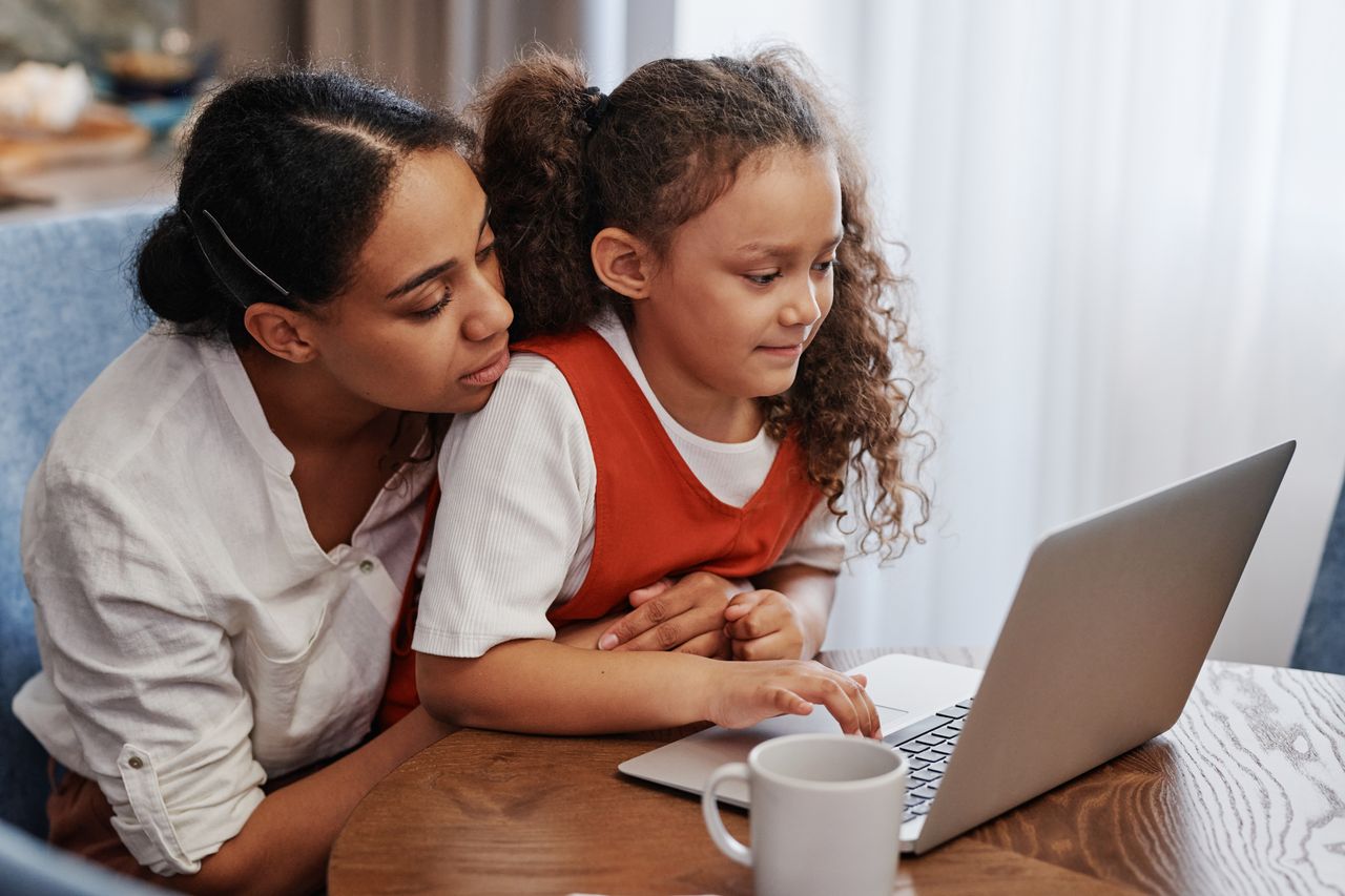 An adult and child sit together at a table looking at a laptop.
