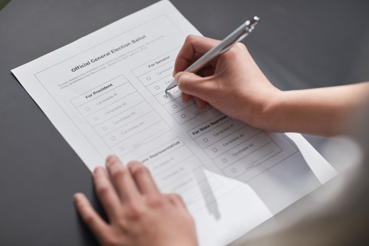 Hands holding a pen over a staged sample election ballot with generic candidate labels.