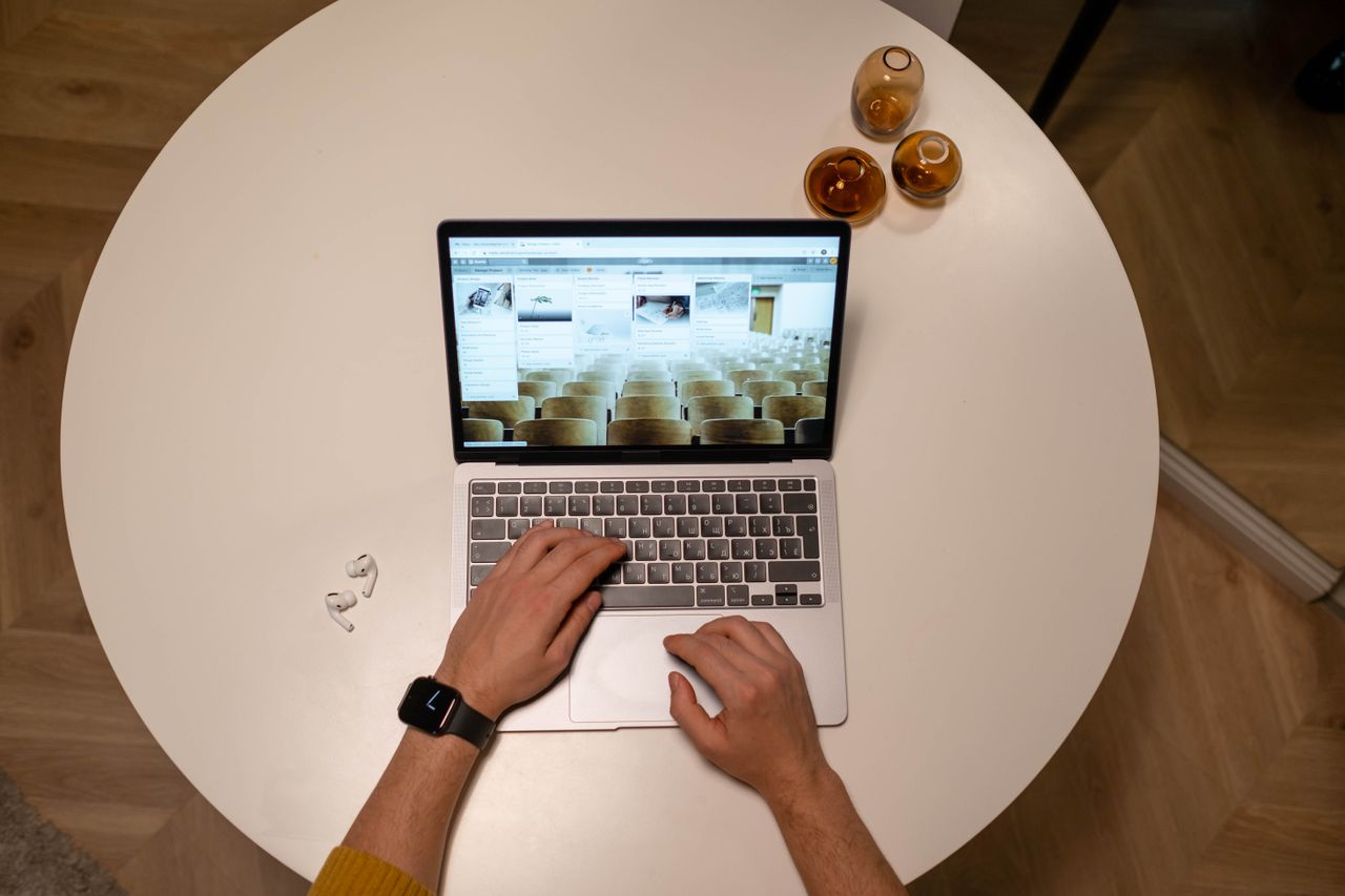Hands using a laptop on a round white table, with earbuds and small amber glass vases nearby.