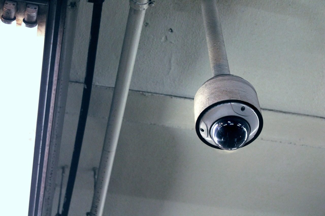 A dome security camera hangs beneath a ceiling beside a window.
