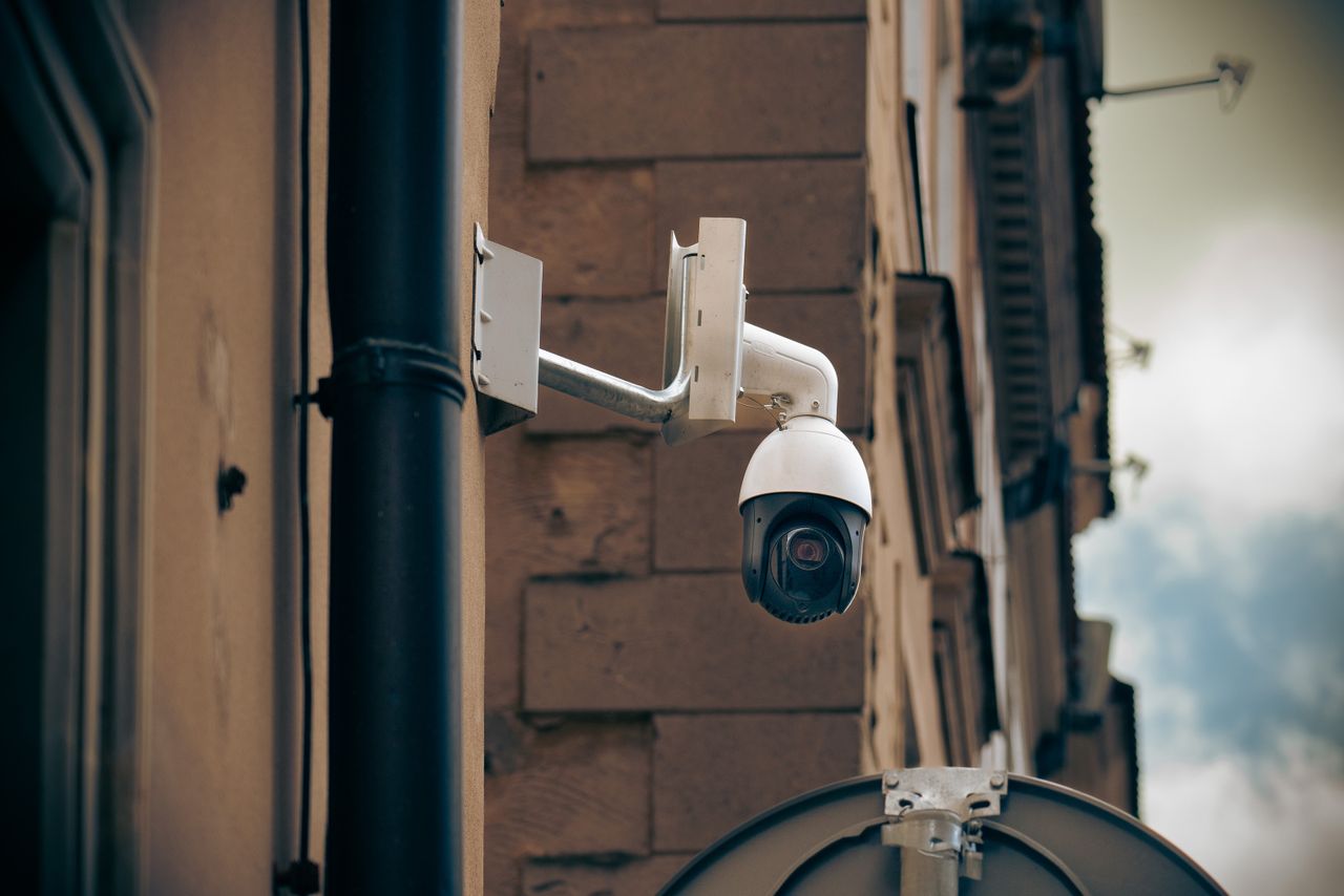 A white security camera with a dark rounded lens housing is mounted on the outside wall of a building.