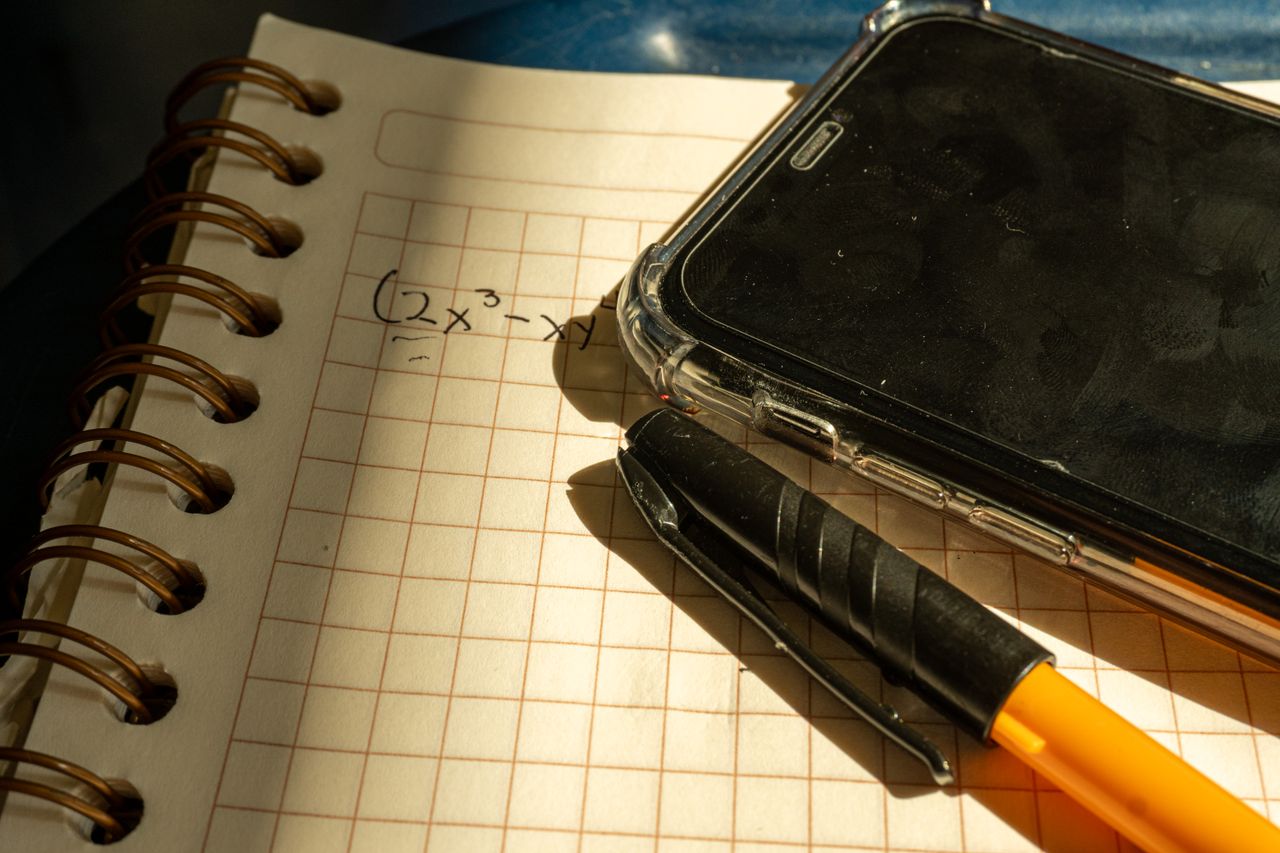 A smartphone and black and yellow pen rest on an open grid notebook with a handwritten math expression.