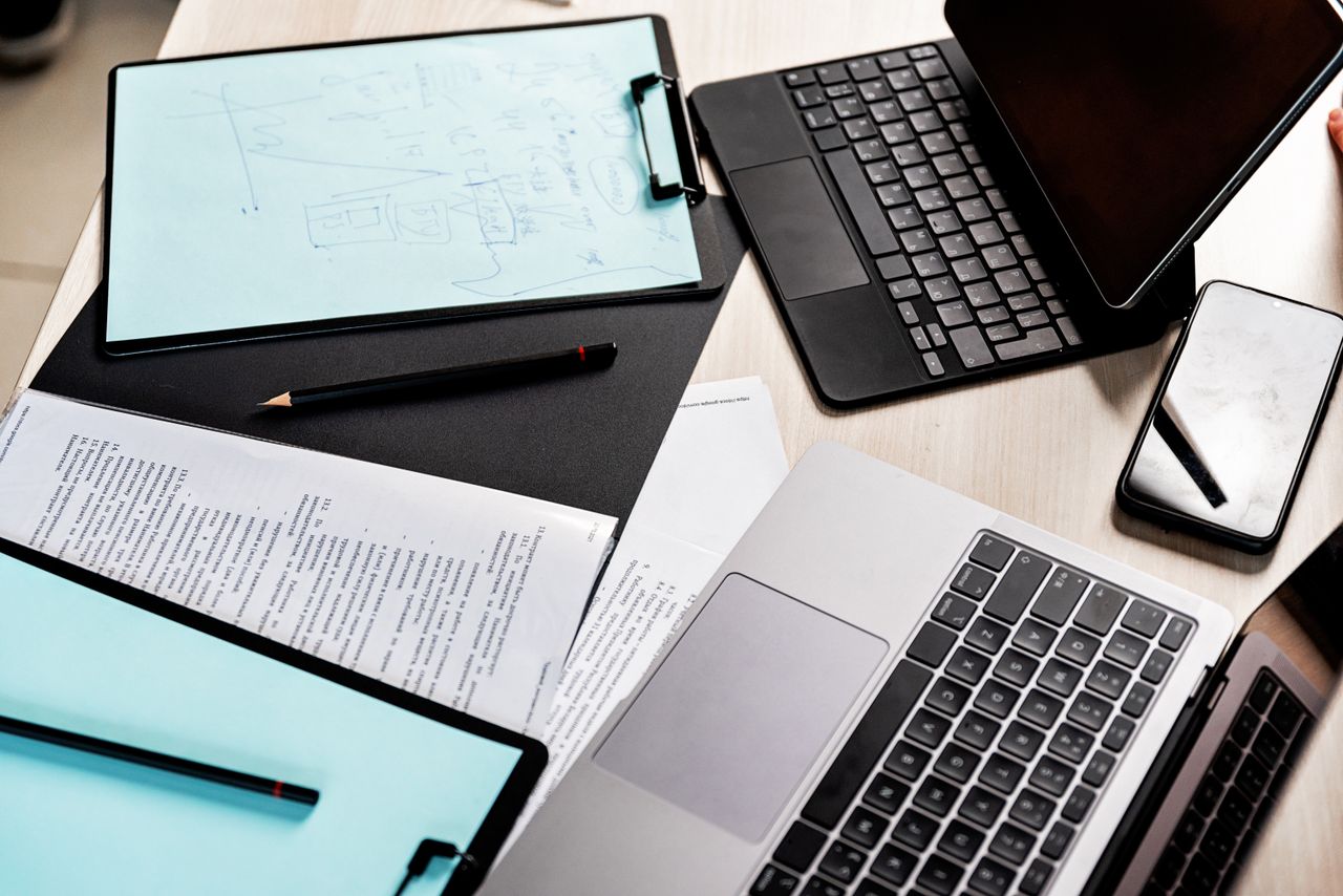 Keyboards, screens, a phone, clipboards and paper documents share a desk.