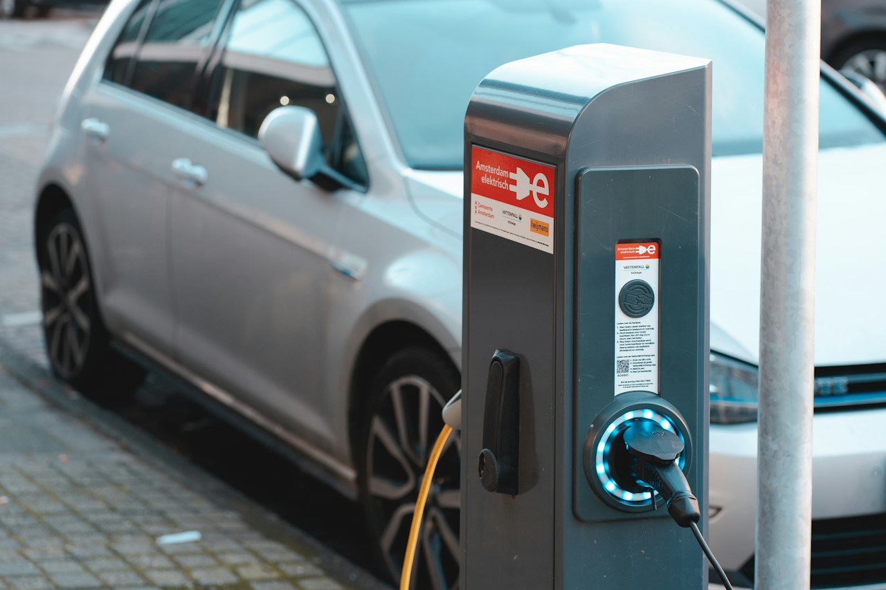 A roadside charging pedestal with a connected cable beside a silver car.