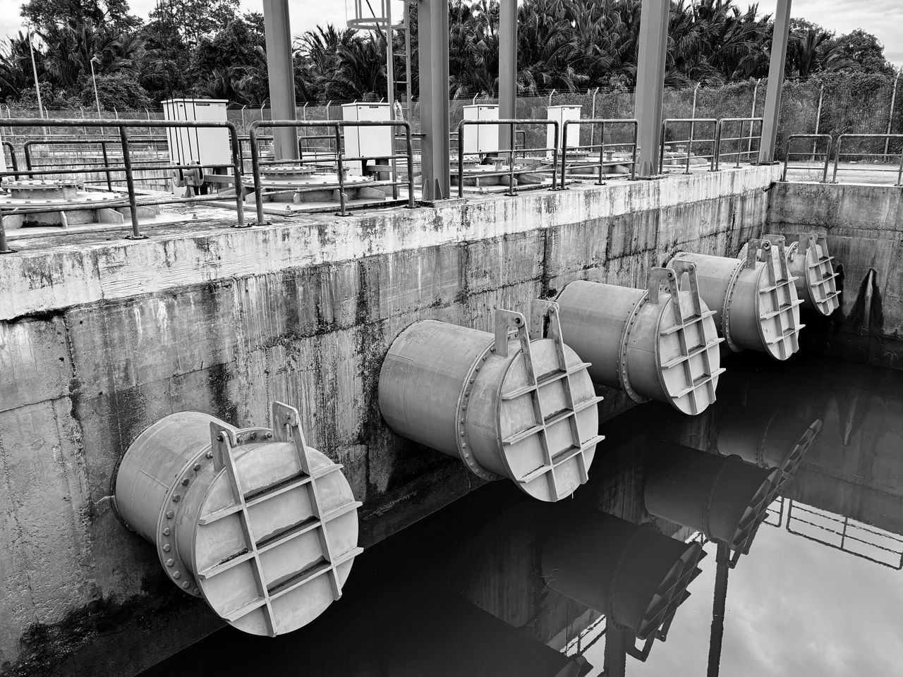 Large covered pipe outlets line a concrete wall above water, with railings and electrical cabinets behind them.