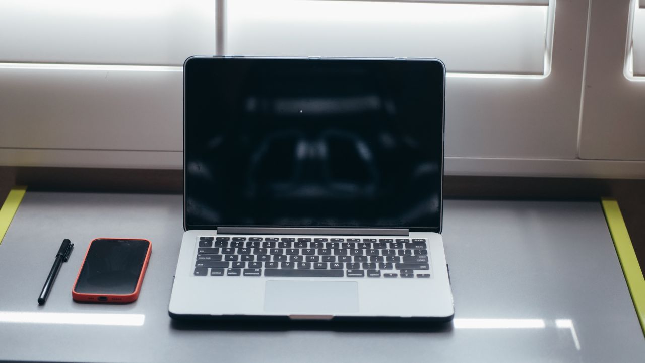 An open laptop with a dark screen beside a smartphone and pen on a desk beneath a window.