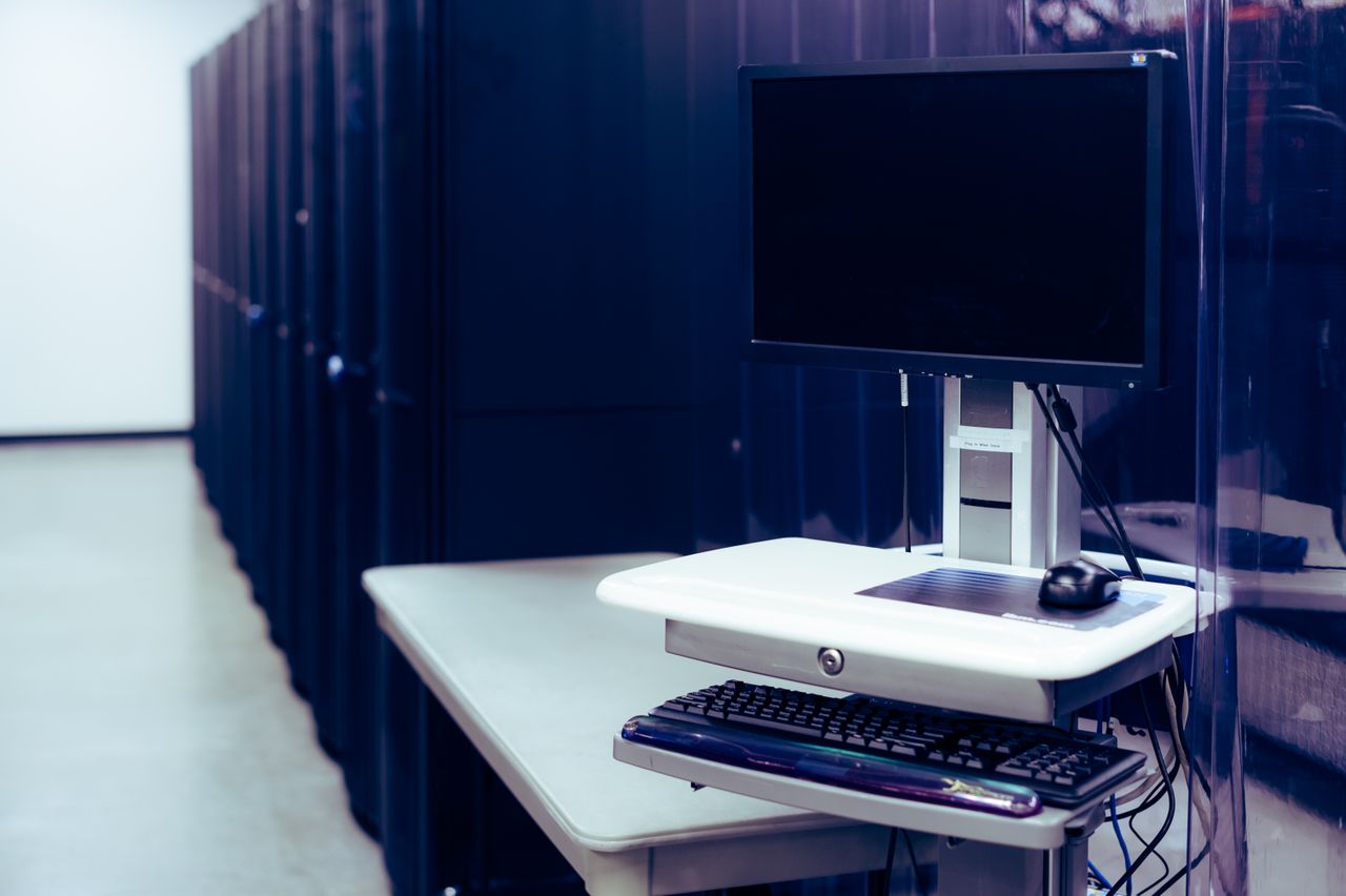 A dark computer monitor, keyboard and mouse on a white workstation beside server cabinets.